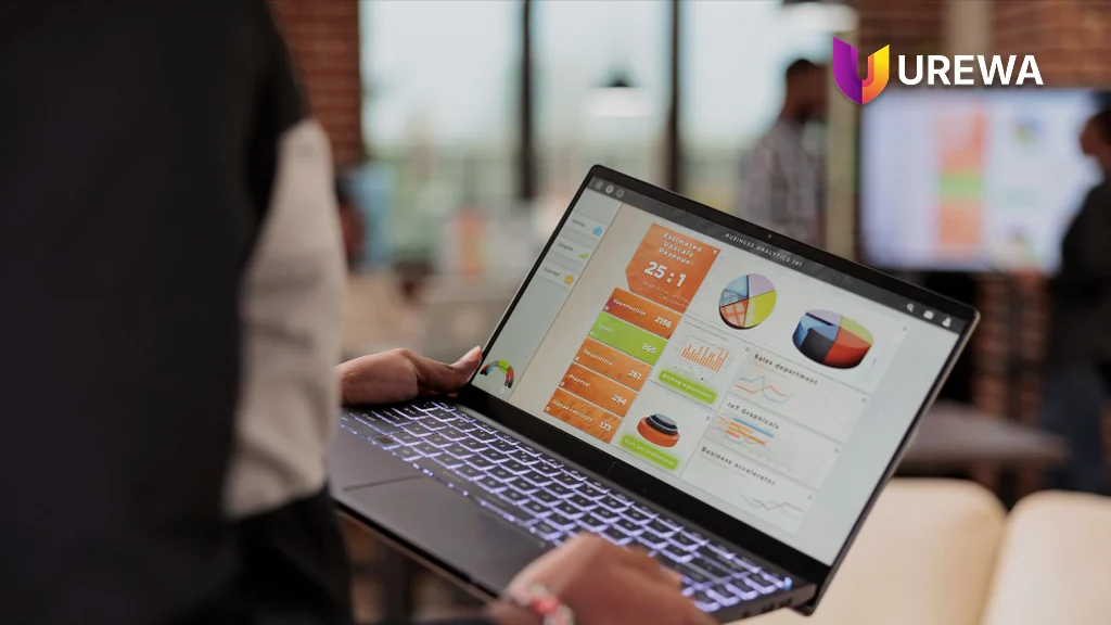 A consultant carrying a laptop showing a Dynamics 365 business analytics dashboard across an office floor during post-go-live hypercare, monitoring operational KPIs while supporting users at their desks