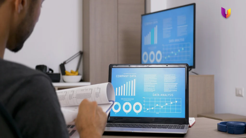 A sales analyst reviewing company data and pipeline charts across a laptop and second screen while working through a printed report, checking conversion and forecast figures
